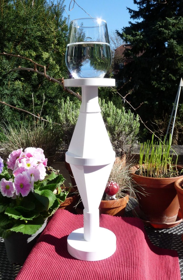 Tisch-Doppelpyramide mit Energetisierungsplatform Eine Tisch-Doppelpyramide mit Energetisierungsplatform und einem Glas Wasser oben drauf auf einem Tisch auf einer Terasse mit Blumen